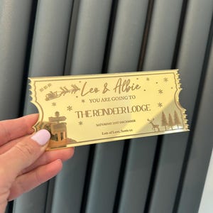 Pode incluir: Bilhete dourado com o texto "Leo & Albie * You are going to * The Reindeer Lodge * Saturday 21st December * Lots of Love, Santa xx" e uma ilustra&ccedil;&atilde;o de uma rena e pinheiros.