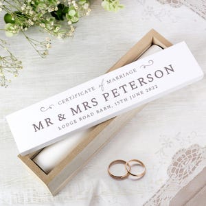 May include: A white wooden box with a lid inscribed with "CERTIFICATE of MARRIAGE MR & MRS PETERSON LODGE ROAD BARN, 15TH JUNE 2022". Two gold wedding rings are in front of the box. The box is on a white surface.