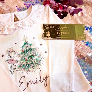 Pode incluir: Uma camisa rosa de manga comprida com gola de renda branca e um design brilhante de &aacute;rvore de Natal e bailarina. A camisa &eacute; personalizada com o nome "Emily". Um bilhete dourado com o texto "Sophie You are going to Lapland" est&aacute; no fundo.