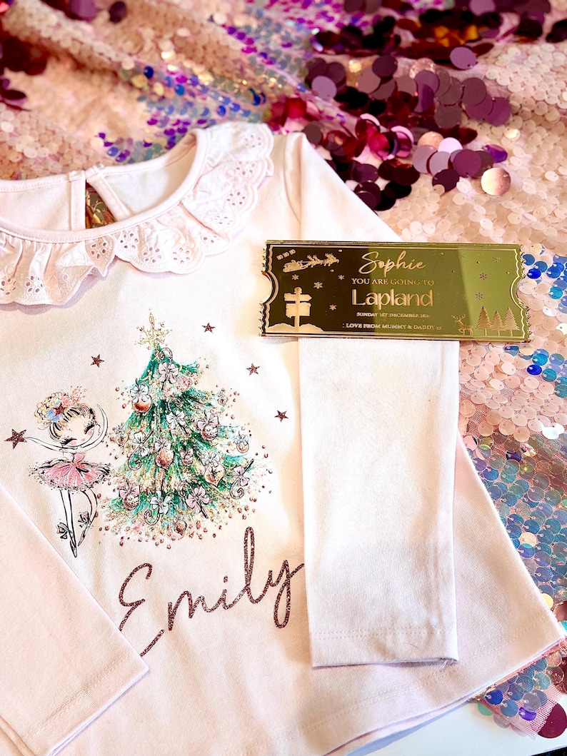 Pode incluir: Uma camisa rosa de manga comprida com gola de renda branca e um design brilhante de &aacute;rvore de Natal e bailarina. A camisa &eacute; personalizada com o nome "Emily". Um bilhete dourado com o texto "Sophie You are going to Lapland" est&aacute; no fundo.