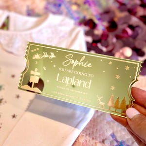 Pode incluir: Um bilhete dourado com glitter com o texto "You are going to Lapland" e o nome "Sophie". O bilhete tem uma rena e &aacute;rvores de Natal.
