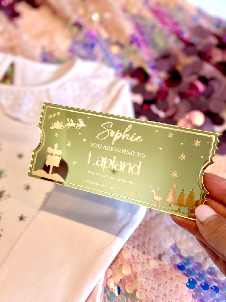 Pode incluir: Um bilhete dourado com glitter com o texto "You are going to Lapland" e o nome "Sophie". O bilhete tem uma rena e &aacute;rvores de Natal.