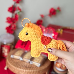 Può includere: Un portachiavi a forma di cavallo all'uncinetto giallo e arancione fatto a mano con una sella rossa. Il cavallo ha un piccolo ciondolo dorato con caratteri cinesi. Il portachiavi è tenuto in mano da una persona, con uno sfondo sfocato di decorazioni rosse.