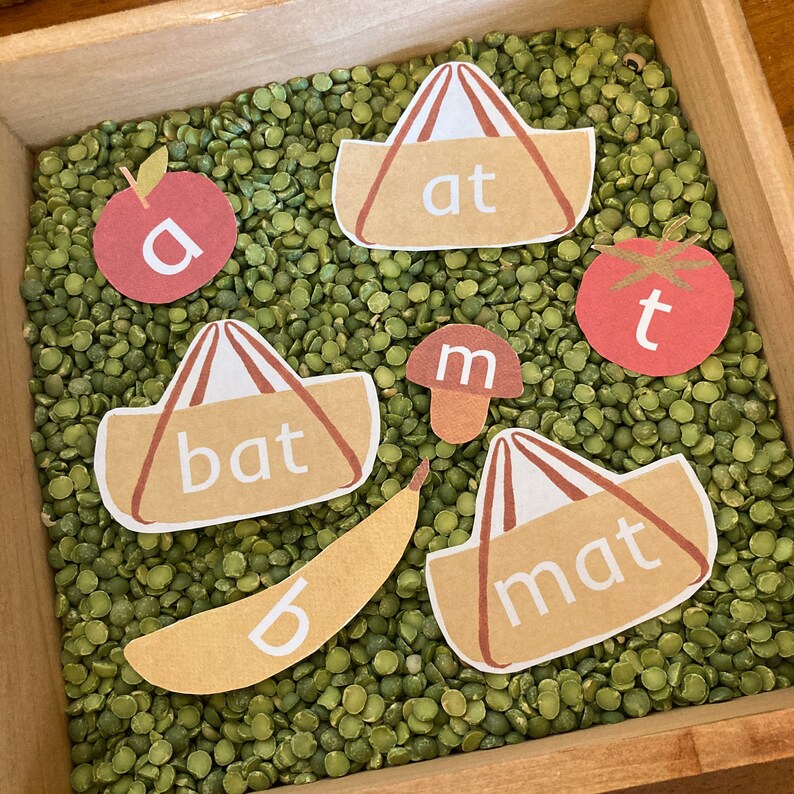 May include: A brown wooden box filled with green split peas.  There are several brown felt bags with the words "at", "bat", "mat" written on them.  There is a red apple with the letter "a" on it, a red tomato with the letter "t" on it, and a brown mushroom with the letter "m" on it.