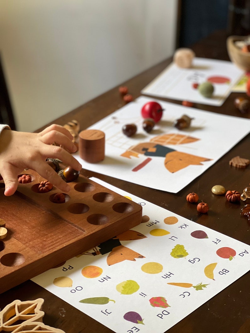 May include: A wooden tray with a grid of holes filled with small wooden balls. A child's hand is reaching into the tray to pick up a ball. There are several printed cards with pictures of fruits and vegetables and the letters Aa, Bb, Cc, Dd, Ee, Ff, Gg, Hh, Ii, Kk, Ll, Mm, Nn, Oo, Pp.