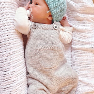 Puede incluir: Un bebé recién nacido con un gorro de punto azul claro, un peto de punto gris claro con dos botones grandes y calcetines grises. El bebé está acostado sobre una manta de punto blanca y tiene un chupete en la boca.