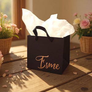 May include: A square black gift bag with black fabric handles and white tissue paper protruding from the top. The front of the bag features the name Esme written in a metallic copper-colored cursive script font.