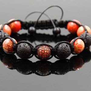 Puede incluir: Un brazalete de cuentas de lava negra y jaspe rojo con una sola cuenta de cristal rojo en el centro. El brazalete está hecho con cordón negro y tiene un cierre de nudo deslizante.