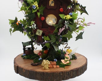 May include: A whimsical fairy garden scene featuring a dark brown cuckoo clock adorned with ivy, flowers, and ladybugs. The clock sits atop a wooden cake stand, surrounded by miniature figurines of a fairy, rabbits, and a well. The scene is illuminated with soft lighting.