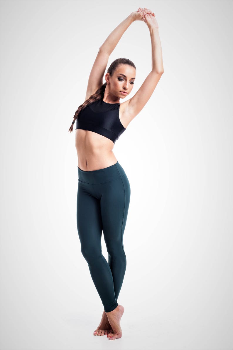 Puede incluir: Una mujer con un sujetador deportivo negro y leggings verde oscuro est&aacute; estirando con los brazos levantados por encima de la cabeza.