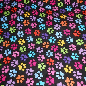 Könnte beinhalten: Ein schwarzer Stoff mit einem sich wiederholenden Muster aus bunten Pfotenabdrücken. Die Pfotenabdrücke sind in verschiedenen Schattierungen von Rosa, Blau, Grün, Orange, Gelb und Lila.