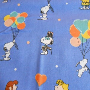 Peut inclure: Tissu bleu avec un motif répété de personnages de Snoopy. Snoopy est représenté dans différentes poses, notamment en tenant des ballons, en portant un chapeau haut de forme et des lunettes, et en volant avec des ballons. L'arrière-plan est bleu clair avec des points blancs.