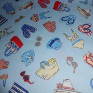 Op de afbeelding: Een blauwe stof met een herhalend patroon van strandartikelen, waaronder zonnebrillen, slippers, strandtassen en badpakken. Het patroon is in verschillende kleuren, waaronder rood, geel, blauw en wit.