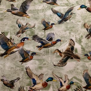 Könnte beinhalten: Stoff mit einem sich wiederholenden Muster von fliegenden Enten in verschiedenen Farben, darunter Braun, Blau und Grün. Der Hintergrund zeigt eine verblasste grüne Landschaft mit Häusern und Bäumen. Die Enten sind im Flug, mit ausgebreiteten Flügeln.