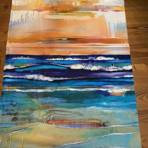 Op de afbeelding: Abstracte stoffen kunst die een kustlandschap uitbeeldt. Het kunstwerk toont een levendige zonsondergang met oranje en gele tinten, die overgaat in een diepblauwe oceaan met witte golven. De onderkant toont een zandstrand met hints van groen en geel.