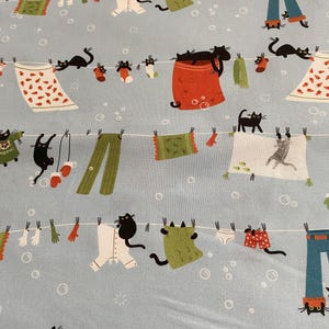 May include: Pale blue fabric featuring a repeating pattern of black cats, clothing, and accessories hanging on washing lines. The clothing includes trousers, shirts, socks, and towels. The cats are in various poses, adding a playful touch to the design.