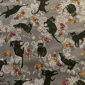 Könnte beinhalten: Grauer Stoff mit einem sich wiederholenden Muster aus schwarzen Katzen in verschiedenen Posen, durchsetzt mit Blumenarrangements in Rosa, Gelb und Lila. Der Hintergrund weist ein subtiles, verziertes Blumenmuster auf.