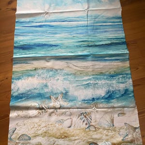 Könnte beinhalten: Ein rechteckiger Stoff mit einer Aquarell-Strandszene. Das Design zeigt einen blauen Himmel mit Wolken, der in Blautöne und Türkis übergeht, die das Meer darstellen. Der untere Teil zeigt einen Sandstrand mit Muscheln und Felsen.
