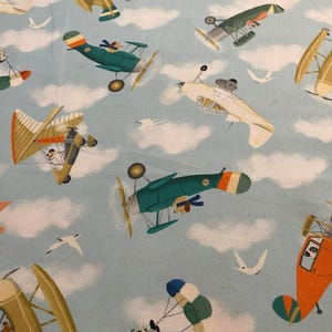 Könnte beinhalten: Hellblauer Stoff mit einem sich wiederholenden Muster aus Oldtimer-Flugzeugen, Wolken und Möwen. Die Doppeldecker sind in verschiedenen Farben gehalten, darunter Grün, Orange und Beige, mit Cartoon-Piloten. Das Design erinnert an ein skurriles Retro-Luftfahrt-Thema.