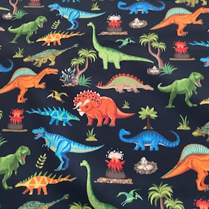 Può includere: Tessuto blu scuro con un motivo ripetuto di dinosauri colorati. I dinosauri sono raffigurati in arancione, verde, blu e giallo. Il design include anche palme, vulcani e uova di dinosauro, con un tema preistorico.