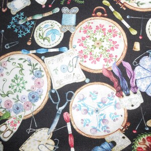 Può includere: Tessuto nero con un motivo ripetitivo di cerchi da ricamo, aghi, forbici e rocchetti di filo. I cerchi da ricamo sono riempiti con disegni floreali colorati.