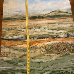 Könnte beinhalten: Stoff mit einem Aquarell-Landschaftsdesign mit sanften Hügeln, Feldern und einem bewölkten Himmel. Die Farbpalette umfasst Grün-, Braun- und Blautöne. Ein gelbes Maßband ist vertikal auf dem Stoff platziert und zeigt die Maße in Zentimetern an.