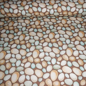 Op de afbeelding: Een bruine stof met een herhalend patroon van witte, beige en blauwe gevlekte eieren. De eieren zijn van verschillende groottes en vormen.