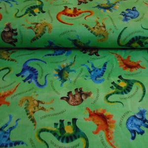Op de afbeelding: Groene stof met een herhalend patroon van kleurrijke dinosaurussen. De dinosaurussen zijn zwart omlijnd en hebben namen eronder geschreven. De namen omvatten Stegosaurus, Triceratops en Tyrannosaurus.