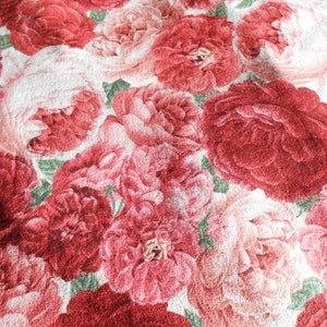 Op de afbeelding: Close-up van een stof met een herhalend bloemenpatroon. Het ontwerp toont grote pioenrozen in tinten rood, roze en wit, met groene accenten. De stof heeft een zachte, getextureerde uitstraling.