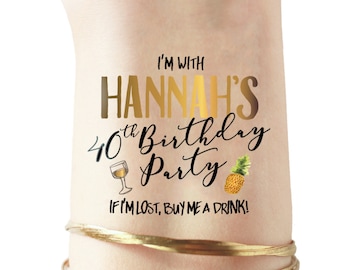 Puede incluir: Tatuaje temporal dorado con el texto "I'm with Hannah's 40th Birthday Party" y un gráfico de una copa de vino y una piña. El texto "If I'm lost, buy me a drink!" está debajo del texto principal.