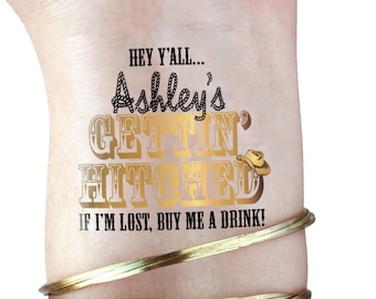 May include: A temporary tattoo with the text "Hey y'all... Ashley's Gettin' Hitched! If I'm lost, buy me a drink!" in black and gold lettering. The text is surrounded by a rope design. A gold cowboy hat is on the right side of the text.