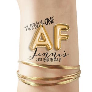 May include: Gold balloon letters spelling "AF" with the text "Twenty-One" and "Jenna's 21st Birthday!" on a person's wrist.