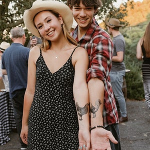 May include: A young woman wearing a black floral dress and a straw cowboy hat stands next to a young man wearing a red and black plaid shirt. They are both smiling and looking at the camera. The man has a temporary tattoo of a rooster on his arm.