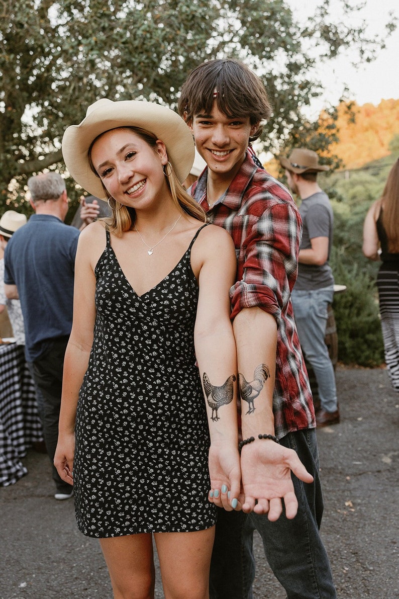 May include: A young woman wearing a black floral dress and a straw cowboy hat stands next to a young man wearing a red and black plaid shirt. They are both smiling and looking at the camera. The man has a temporary tattoo of a rooster on his arm.