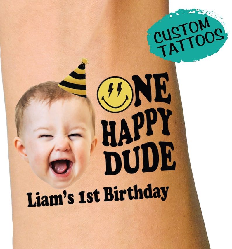 May include: A black and yellow temporary tattoo with a smiley face, a party hat, and the text 'One Happy Dude' with the name 'Liam's 1st Birthday' below. The tattoo is on a person's arm.