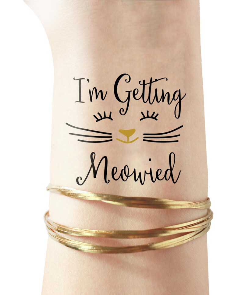 Puede incluir: Un tatuaje temporal con el texto "I'm Getting Meowied" y una cara de gato de dibujos animados con pesta&ntilde;as y una nariz dorada.