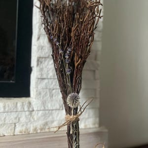 Puede incluir: Un arreglo floral seco con un manojo de ramitas marrones, acentuado con pequeñas flores moradas y un cardo. El arreglo está atado con hilo de cáñamo, creando una estética rústica y natural. Perfecto para la decoración del hogar.