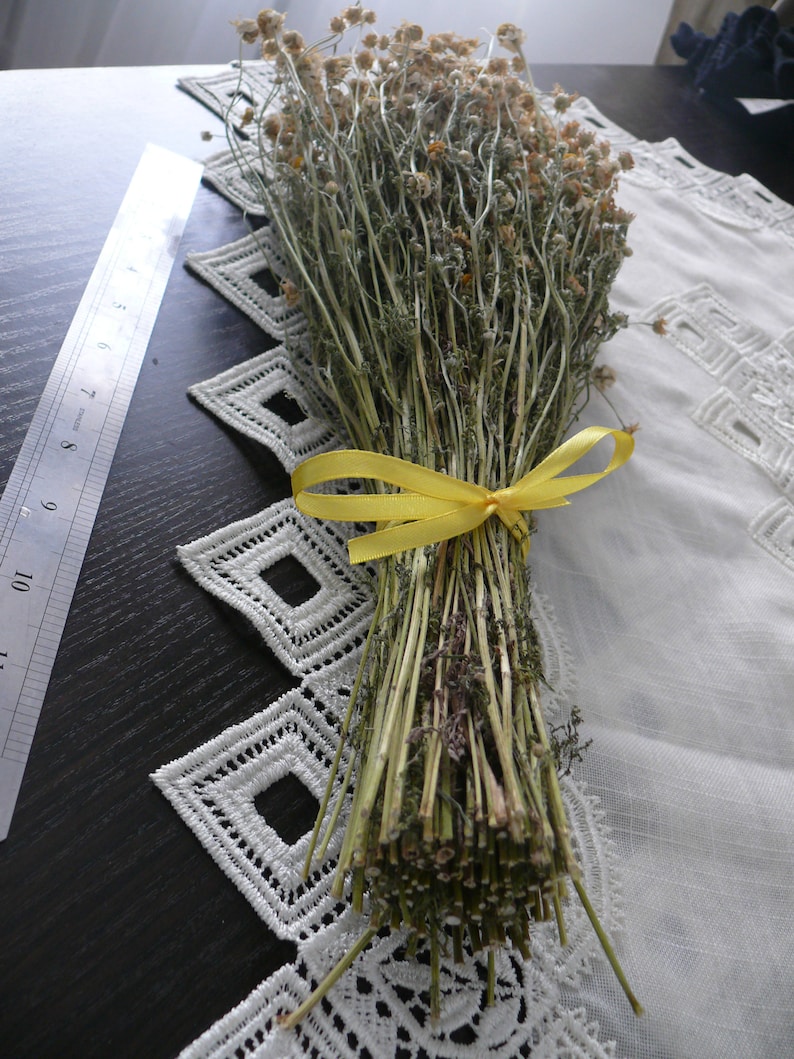 Puede incluir: Un manojo de flores de manzanilla secas atadas con una cinta amarilla. Las flores est&aacute;n dispuestas sobre un mantel de encaje blanco.