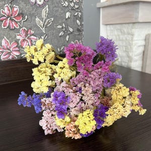 May include: A vibrant bouquet of dried flowers in shades of yellow, purple, pink, and blue. The flowers are arranged on a dark brown wooden surface, creating a colorful and natural display. The bouquet is a mix of textures and hues.