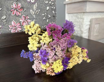 May include: A vibrant bouquet of dried flowers in shades of yellow, purple, pink, and blue. The flowers are arranged on a dark brown wooden surface, creating a colorful and natural display. The bouquet is a mix of textures and hues.