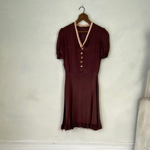 May include: A maroon vintage dress with a white collar and button fastening. The dress has short sleeves and a frilled hem. The dress is hanging on a wooden hanger.