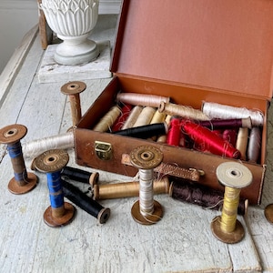 Op de afbeelding: Een vintage bruine leren naaibox met een assortiment garenrollen in verschillende kleuren, waaronder rood, wit en beige. De spoelen zijn gerangschikt in de doos en op een witte houten tafel.