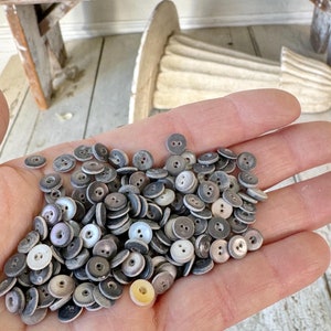 May include: A hand holding a large pile of vintage, gray and white, two-hole buttons. The buttons are made of a smooth, pearly material.