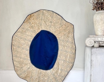 May include: A large, irregularly shaped wall hanging with a blue center and a patterned, cream-colored border. The border has a grid-like design with blue lines. The hanging is mounted on a light gray wall.