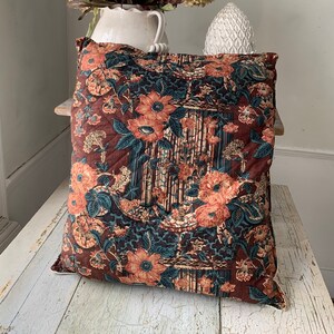 May include: A brown and teal floral patterned throw pillow with a white border. The pillow is sitting on a white wooden surface.