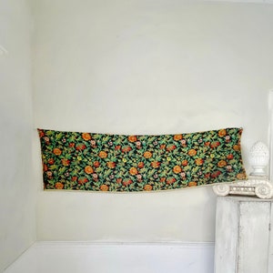 May include: A vintage floral tapestry wall hanging with a dark green background and orange, yellow, and red flowers. The tapestry is hanging on a white wall in a room with white trim and a white painted cabinet.