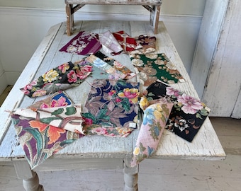 May include: A collection of vintage fabric swatches with floral and geometric patterns in various colors, including red, green, blue, and pink, displayed on a white wooden table with a small wooden stool and a white decorative pineapple finial.