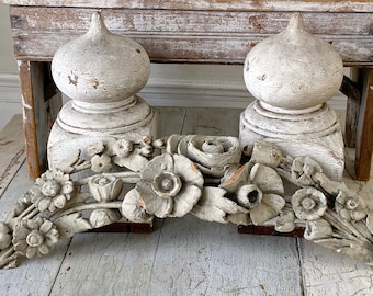 May include: Two white painted wooden finials with a carved floral garland. The finials are shaped like a dome with a pointed top. The garland is made of flowers and leaves. The garland is in a light grey colour.