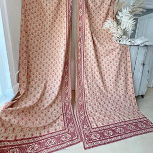 May include: A pair of cream-coloured curtains with a red floral pattern and a red border. The curtains have a vintage look and are made of a lightweight fabric.