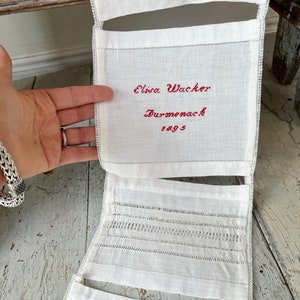 以下が含まれることがあります： 赤い刺繍で「Elina Wacker Durmenach 1893」と刺繍された白いリネン製のサンプラー。サンプラーは、さまざまなレース模様のセクションで構成されています。